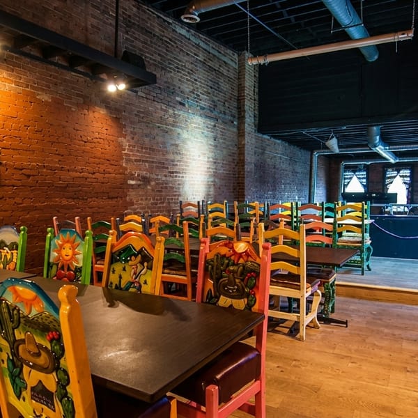 Restaurant interior with colorful, hand-painted chairs at dark wooden tables against a brick wall.
