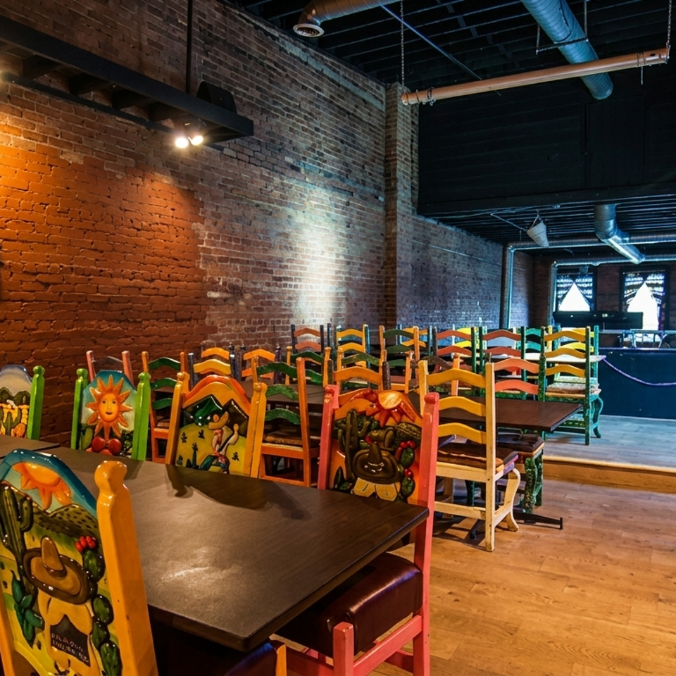 Restaurant interior with colorful, hand-painted chairs at dark wooden tables against a brick wall.