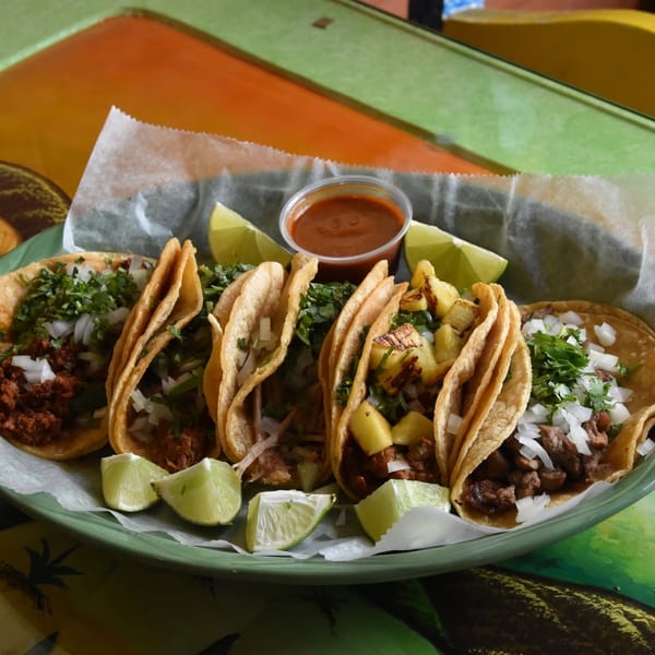 Plate of assorted Mexican tacos topped with cilantro and onions, served with grilled pineapple, lime wedges, and red salsa.
