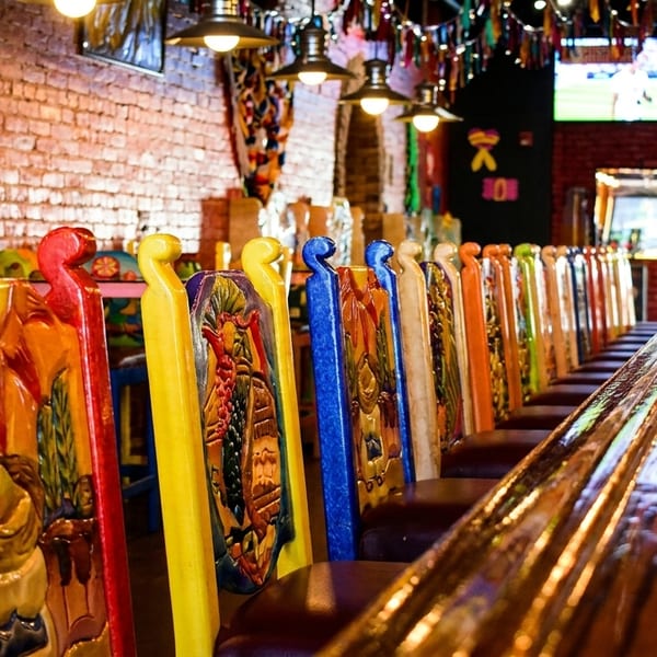 Colorful, carved wooden bar stools line a glossy bar top in a vibrant Mexican restaurant.