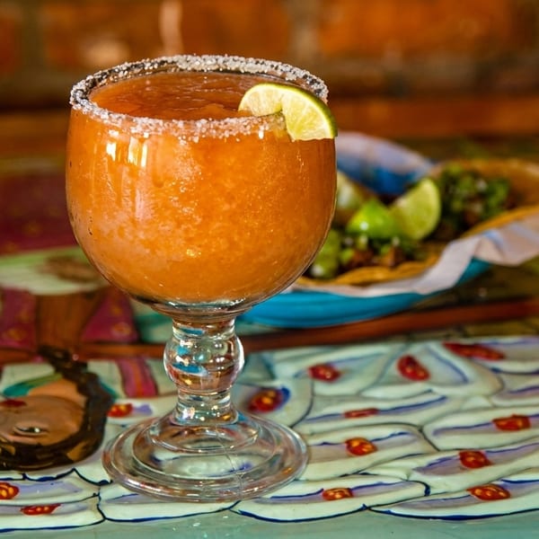 A vibrant orange frozen drink with a lime wedge and salted rim sits on a colorful, patterned table.
