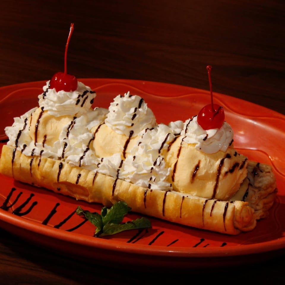Banana split with vanilla ice cream, whipped cream, cherries, and chocolate drizzle on an orange plate.