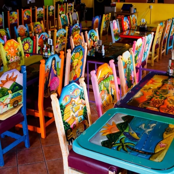 Vibrant, hand-painted chairs and tables fill a colorful Mexican restaurant dining room.