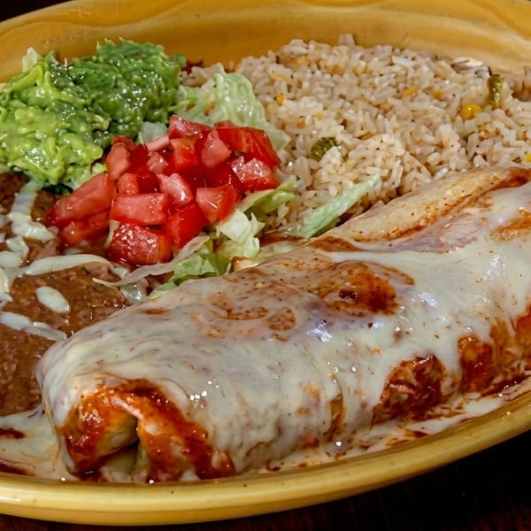 A large burrito covered in melted cheese and red sauce, served with rice, refried beans, guacamole, and pico de gallo.