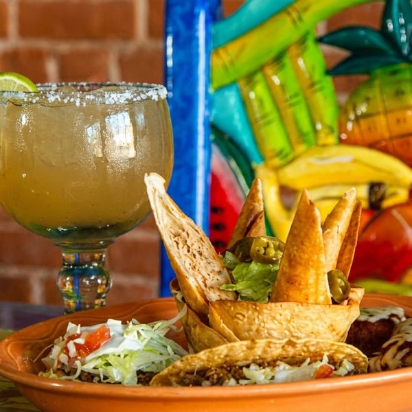 A margarita with a lime wedge and a plate of tacos, taquitos, and other Mexican appetizers.